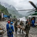 Rescue workers in high-visibility gear pulling a survivor from a dark, mud-caked tunnel entrance in a mountainous region of Nepal.