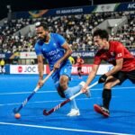An intense action shot of a professional field hockey match at the Asian Games with players competing for the ball on a blue synthetic pitch.