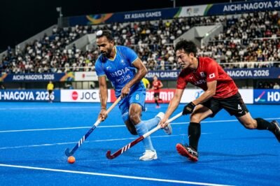 An intense action shot of a professional field hockey match at the Asian Games with players competing for the ball on a blue synthetic pitch.