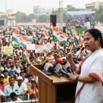West Bengal CM Mamata Banerjee gestures emphatically during a political rally in Kolkata, surrounded by supporters and party flags.