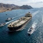 A military patrol boat guarding the narrow shipping lanes of the Strait of Hormuz with an oil tanker in the background.