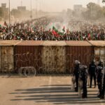 A massive crowd of protesters holding green and red flags faces off against police barriers and shipping containers on a main highway leading to Islamabad.