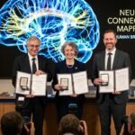 Three Nobel laureates standing in front of a digital display showing neural pathways illuminated by blue and yellow light.