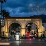 A wide-angle cinematic photo of the iconic Paramount Pictures studio gates with a digital Skydance logo overlay, symbolizing the merger of legacy and modern technology.