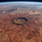 A high-altitude NASA satellite image of the Tnorala impact crater in Australia, resembling a giant red spider against the desert floor.