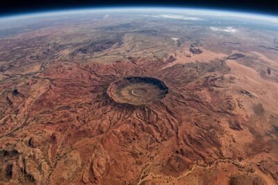A high-altitude NASA satellite image of the Tnorala impact crater in Australia, resembling a giant red spider against the desert floor.